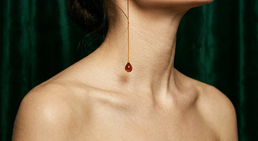 Editorial profile portrait of a woman with luminous skin and a suspended droplet of black seed oil mid-fall against deep emerald velvet